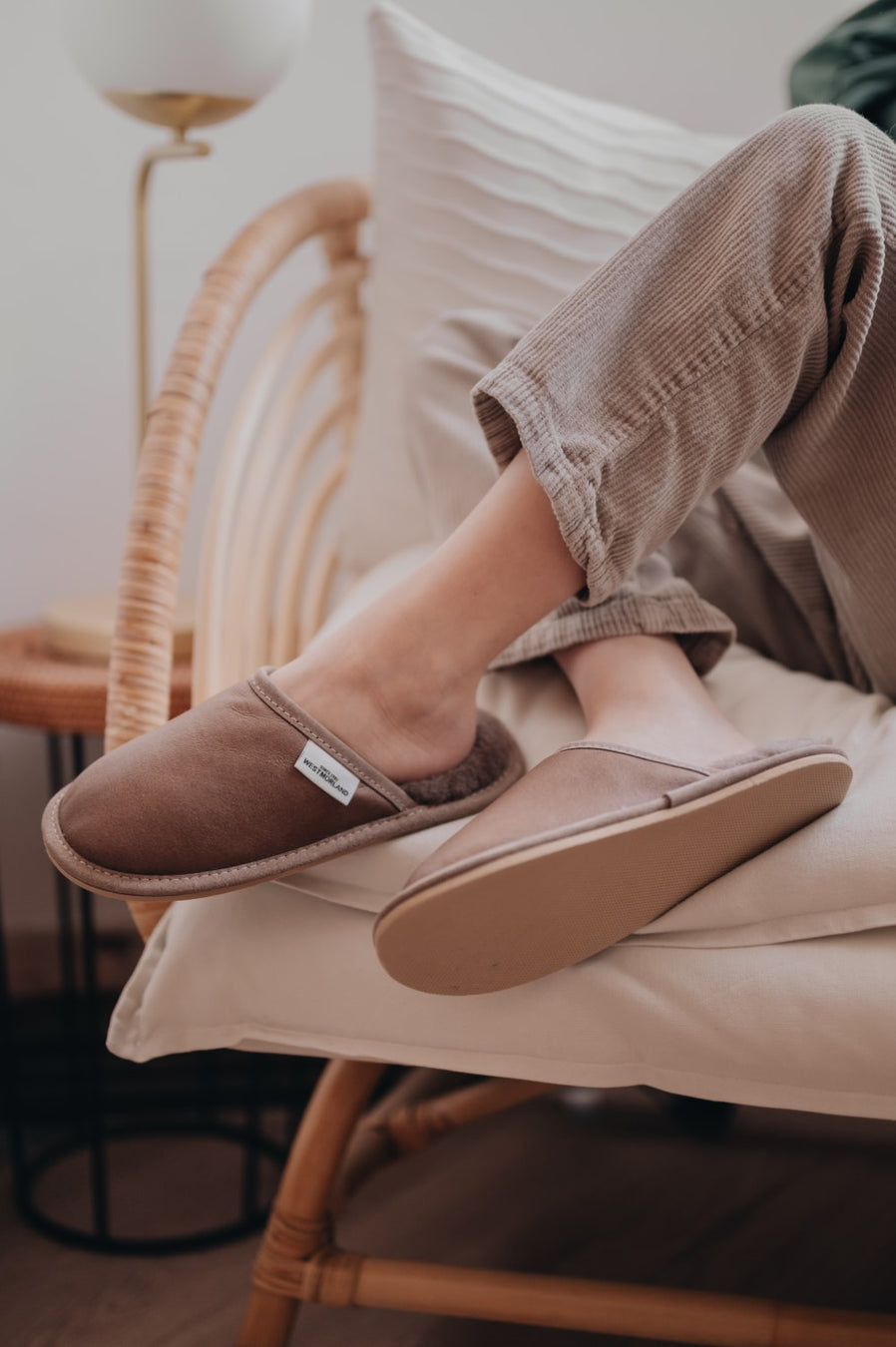 Slip on sheepskin slippers in a brown/grey colour, worn by model lounging in a chair