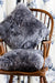 Icelandic Sheepskin Cushion in Grey, on wooden chair against floral background.